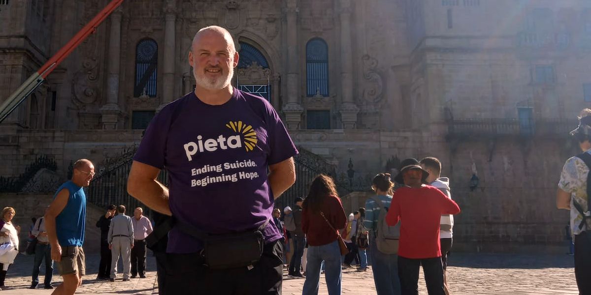 A man in a purple "Pieta" t-shirt stands smiling in front of a historical building, surrounded by a few people on a sunny day.